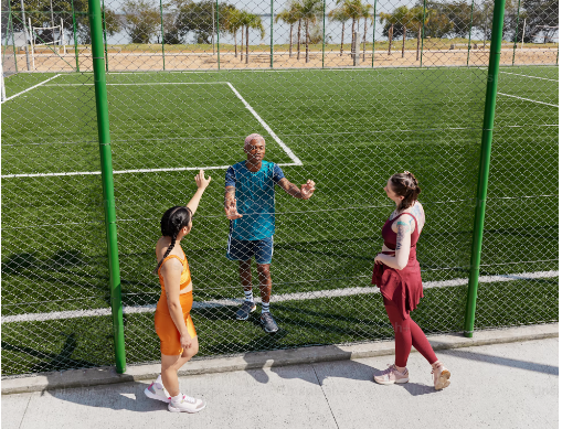 Drei Menschen am Rand eines Fußballfeldes sehen sich durch einen Zaun hindurch an